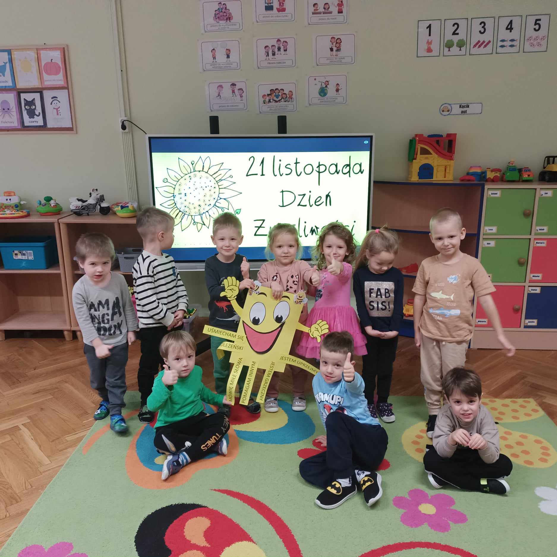 grupa dzieci przedstawia kwadratową postać z żółtej kartki z uśmiechem w tle ekran z napisem dzień życzliwości