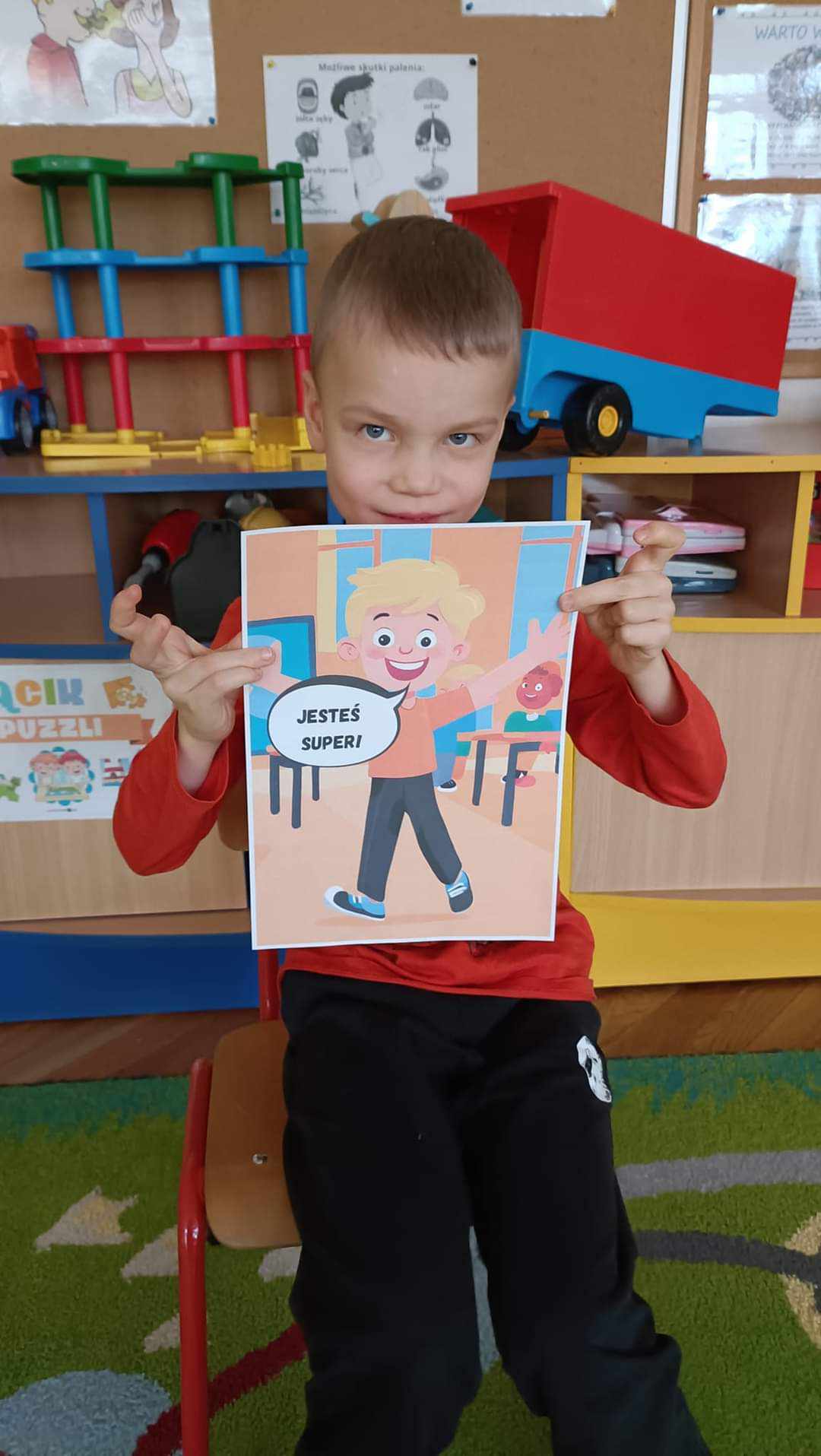 sala przedszkolna chłopiec siedzi na krześle przedstawia kartę z napisem jesteś super