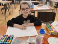 dziewczynka przy stole przed nią biała kartka pisaki farby kredki