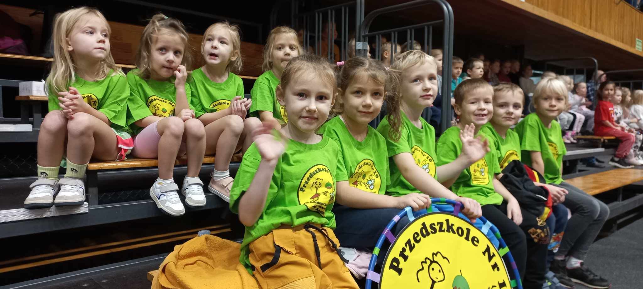 hala sportowa dzieci w zielonych koszulkach siedzą na trybunach trzymają logo przedszkola nr 10 z oddziałami Integracyjnymi Leszczynowa Górka w Koninie