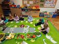 dzieci siedzą na dywanie na środku misy z owocami i warzywami