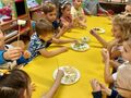dzieci siedzą przy żółtym stole mają wykałaczki na które nadziewają pokrojone owoce