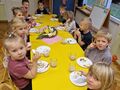 przy żółtych stolikach siedzą dzieci na środku stołu stoi taca z owocami i warzywami. dzieci jedzą kanapki z rzodkiewką i papryką