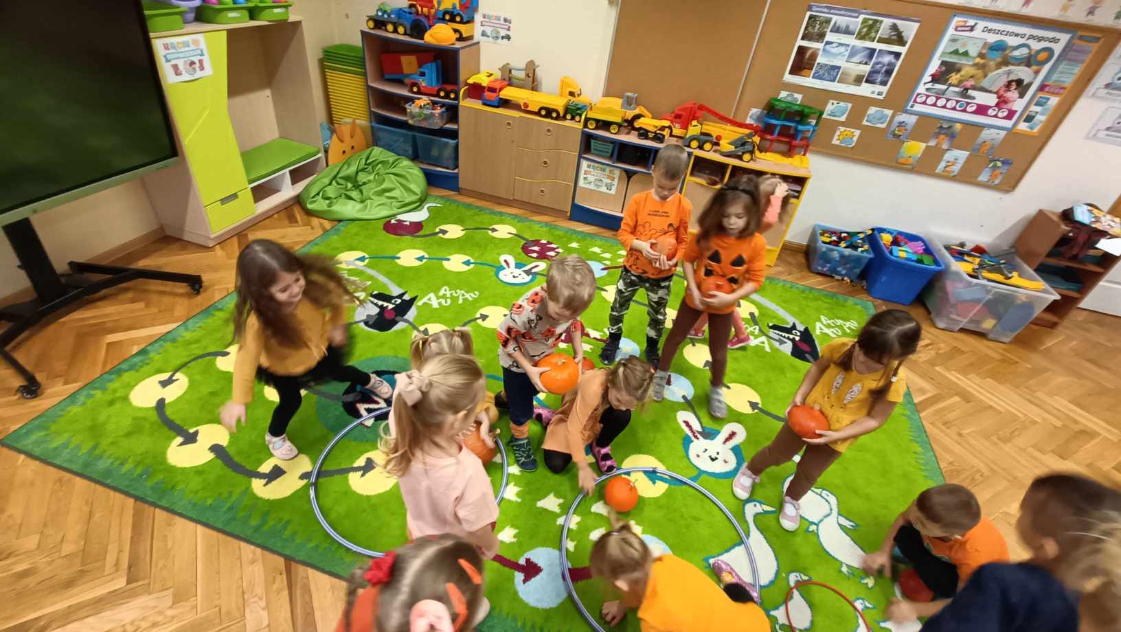 grupa dzieci w przedszkolu ubrana na pomarańczowo wkłada dynie do kół hula hop