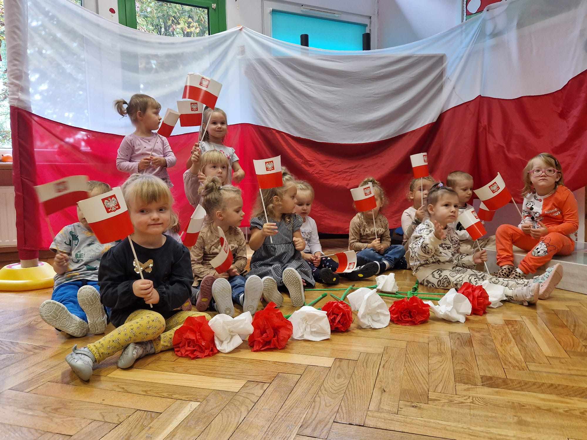 dzieci siedzą na tle flagi Polski trzymają w rękach flagi przed nimi leżą kwiaty białe i czerwone 