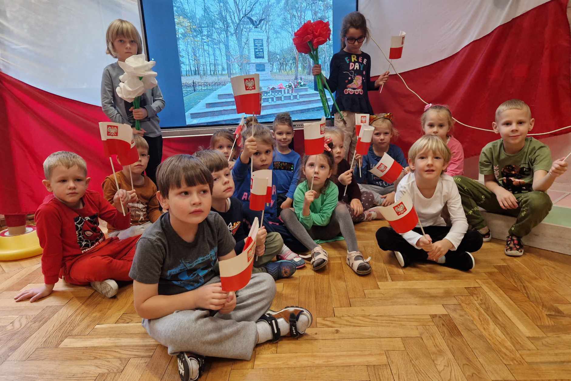 dzieci siedzą przed monitorem wyświetlony pomnik bohaterów w tle flaga Polski 