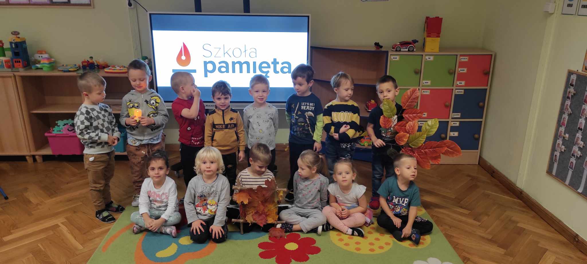 dzieci stoją na tle ekranu multimedialnego z napisem szkoła pamięta, kilkoro dzieci trzyma świeczkę 