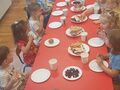 Dzieci siedzą przy czerwonych stolikach i jedzą słodkości