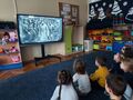 Dzieci siedzą przed ekranem multimedialnym oglądają film o Powstaniu Wielkopolskim