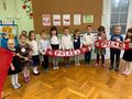 Grupa dzieci stoi trzymając w rękach szaliki z napisem Polska oraz dużą flagę Polski