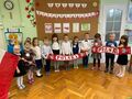 Grupa dzieci ubranych w stroje galowe trzyma 2 szaliki z napisem Polska oraz biało czerwoną flagę