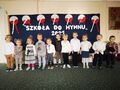 Grupa dzieci śpiewa hymn. Za nimi biały napis na granatowym tle Szkoła do hymnu 2021
