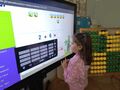 dziewczynka stoi przed ekranem multimedialnym rozwiązuje przykład z dodawania